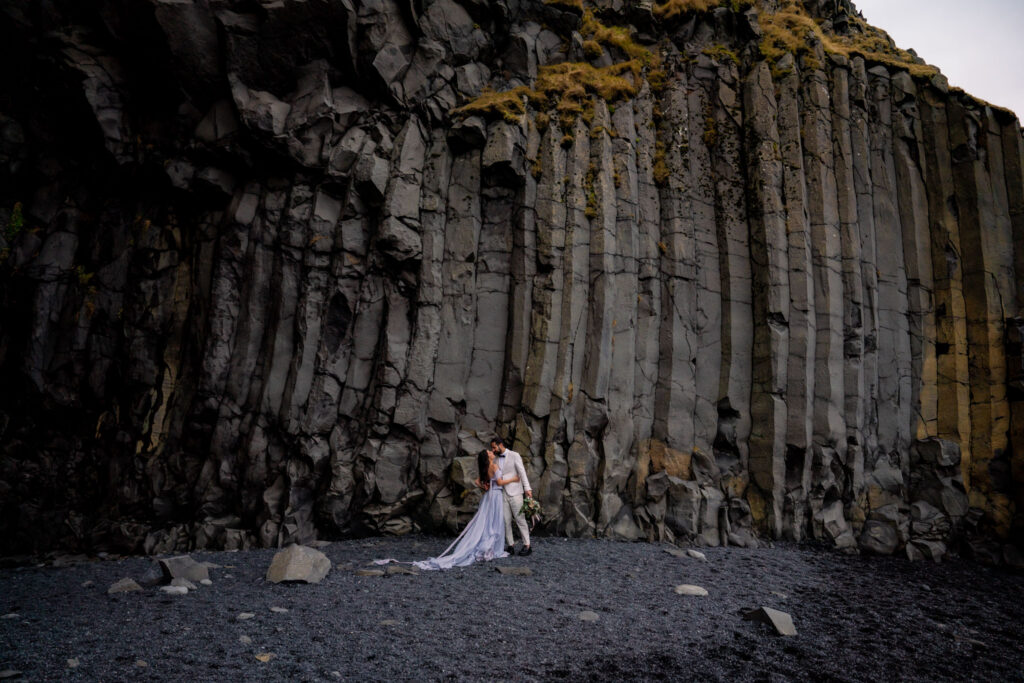 Bride and groom standing together beneath dramatic basalt cliffs during their Iceland black sand beach elopement adventure.