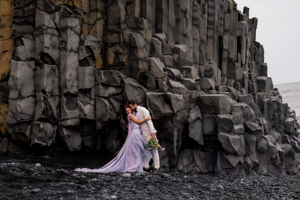 Bride and groom standing beside towering basalt cliffs during their Iceland black sand beach elopement at Reynisfjara.