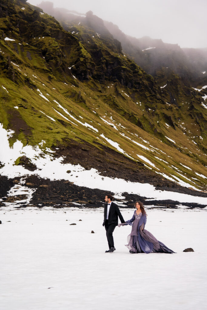 Couple walking together across a snowy Iceland glacier landscape during their adventurous Iceland elopement surrounded by volcanic mountains.