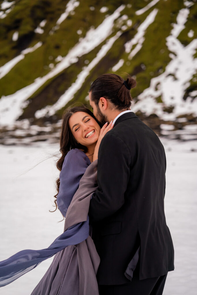 Bride smiling while embracing groom during a Katla ice cave elopement in Iceland with snowy volcanic mountains in the background.