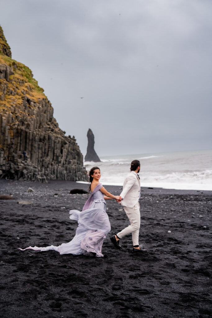 Bride and groom running hand in hand on Reynisfjara black sand beach during their adventurous Iceland elopement with ocean waves in the background.